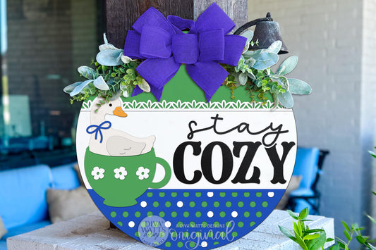Digital SVG file of a round door hanger featuring a goose sitting inside a teacup with “stay cozy” text. This instant download laser cut design is ideal for cottagecore and grandma core DIY décor. The digital file allows you to create your own cozy porch or entryway sign using a laser machine, with layered elements designed for easy cutting, painting, and assembly.